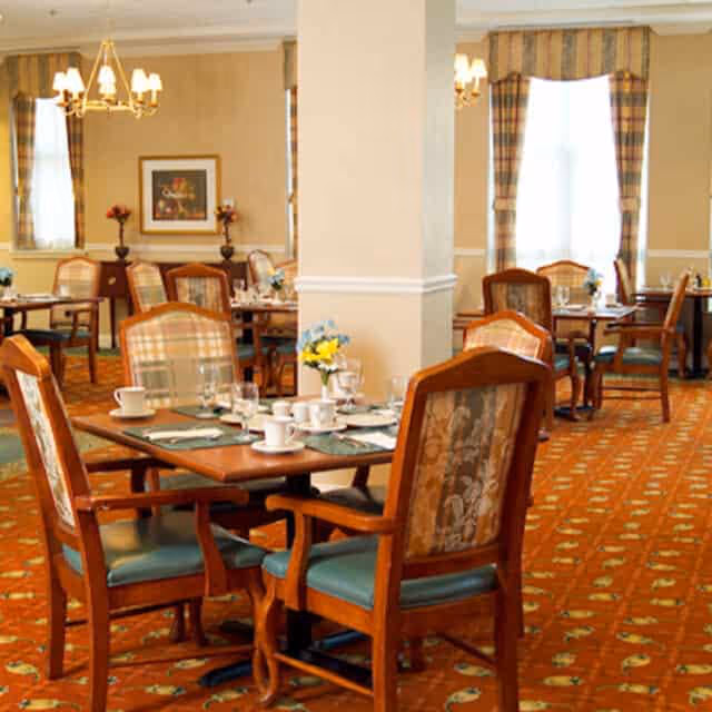 A warmly lit dining room with multiple wooden tables and chairs arranged neatly. Each table is set with white cups, glasses, and silverware. The room features patterned carpet, beige walls, framed artwork, and large windows with striped curtains allowing natural light to enter.