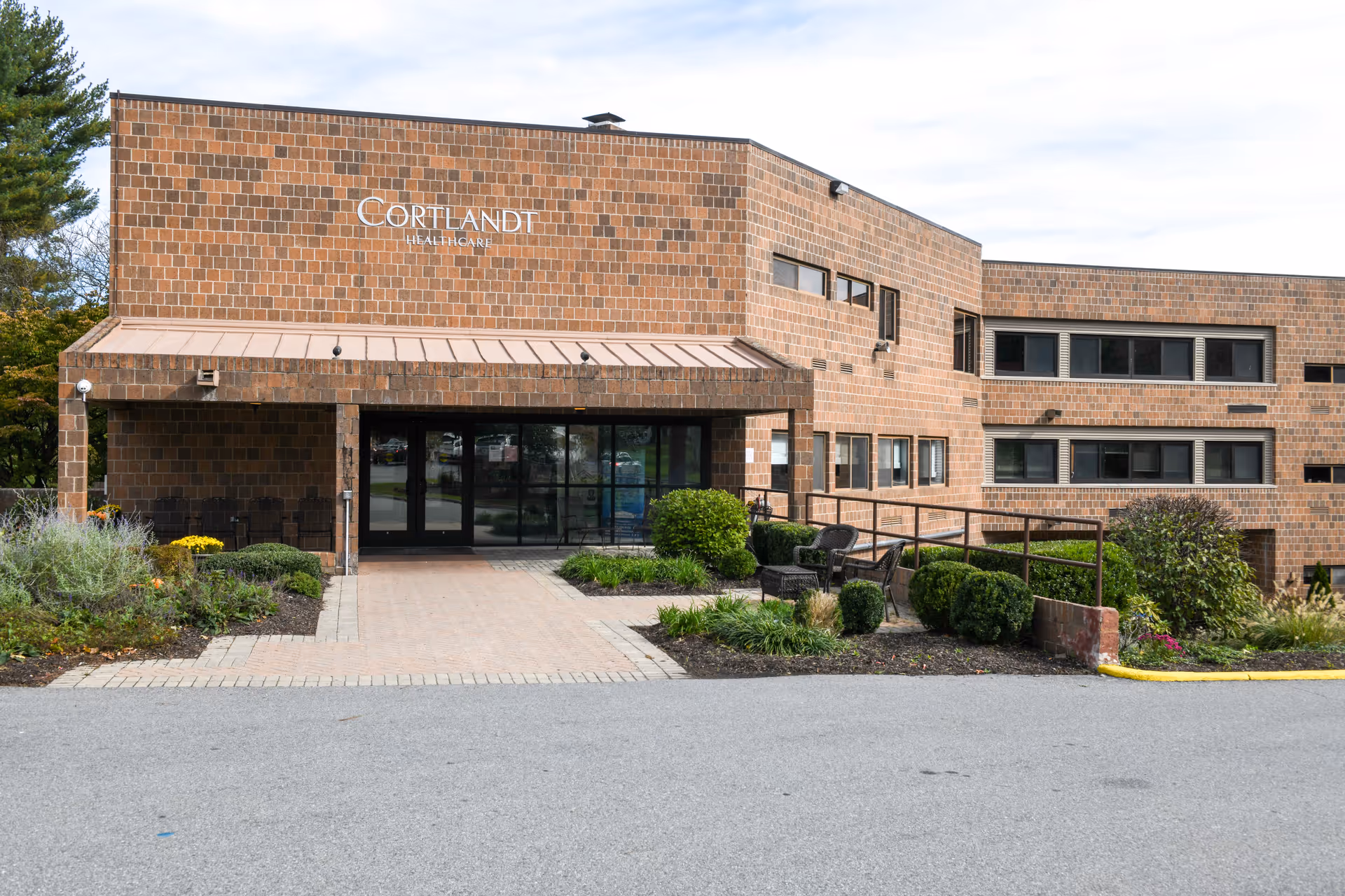Exterior view of Cortlandt Healthcare building with a brick facade, entrance with glass doors, a small garden with bushes and flowers, and a paved driveway in front.