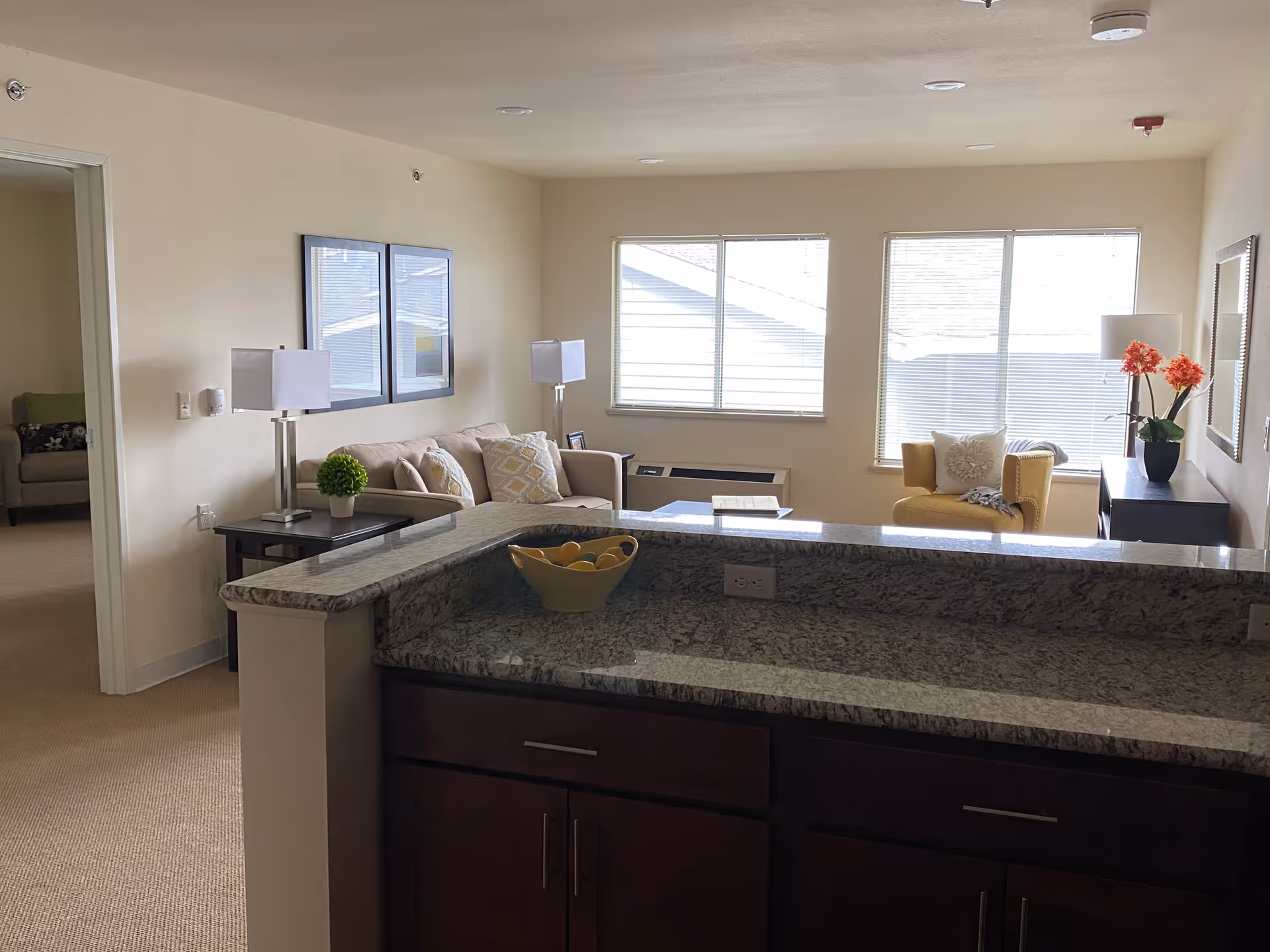 Interior view of a senior living facility's common area featuring a granite countertop with a bowl of fruit, a beige sofa with decorative pillows, a yellow armchair, two table lamps, framed artwork on the wall, and large windows letting in natural light.