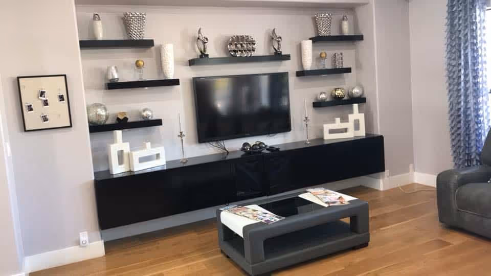 Modern living room with a wall-mounted flat screen TV above a long black floating cabinet. Several black floating shelves hold decorative items such as vases, sculptures, and candles. A small black coffee table with magazines is in front of the cabinet, and part of a gray armchair and blue patterned curtains are visible on the right. The floor is wooden.