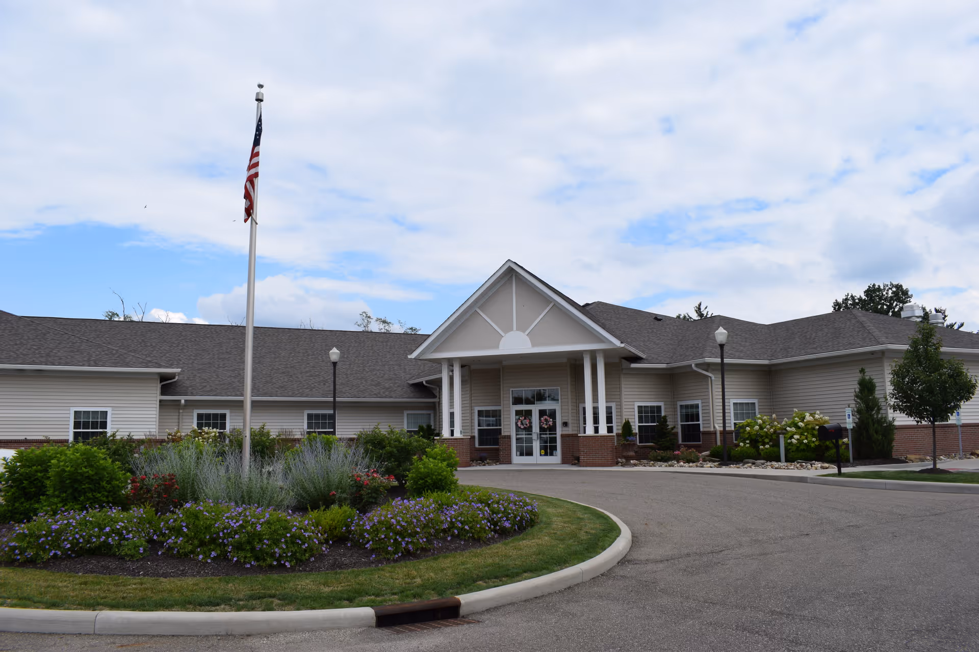 Exterior view of Stone Creek Assisted Living and Memory Care building with a circular driveway, landscaped garden with flowers and shrubs, an American flag on a flagpole, and a partly cloudy sky.