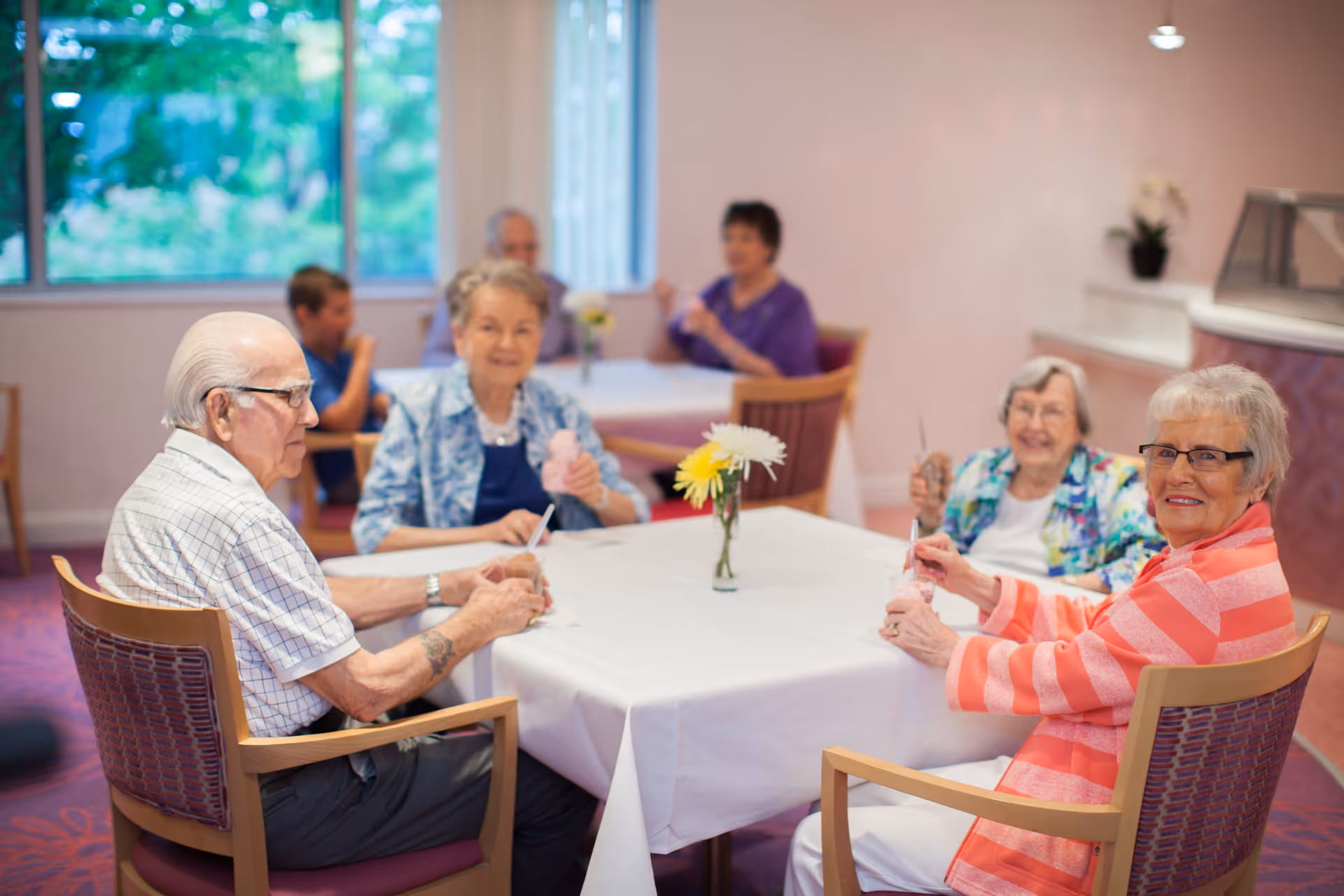 A group of elderly people sitting around a table in a dining area, enjoying ice cream. The room has large windows with a view of greenery outside, and there are flowers in a vase on the table.