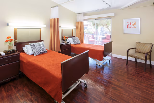 A bright and clean bedroom in a healthcare facility featuring two hospital-style beds with orange bedspreads and patterned pillows. Each bed has a wooden headboard and footboard, and there are nightstands with flowers and books between the beds. A large window with curtains lets in natural light, and a single upholstered chair is placed against the wall. The room has wooden flooring and light-colored walls with a framed artwork.