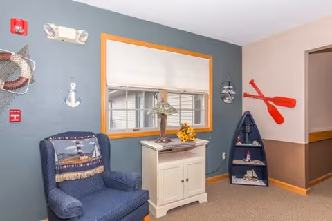 A cozy corner of a living room or common area with a blue upholstered armchair featuring a lighthouse-themed throw blanket. Next to the chair is a small white cabinet with a decorative lamp and a small flower arrangement on top. The wall behind is painted in two tones, with a window framed in wood. Nautical-themed decorations, including a hanging anchor, a fishing net with a starfish, a small boat-shaped shelf with miniature boats, and two red oars mounted on the wall, add to the decor.