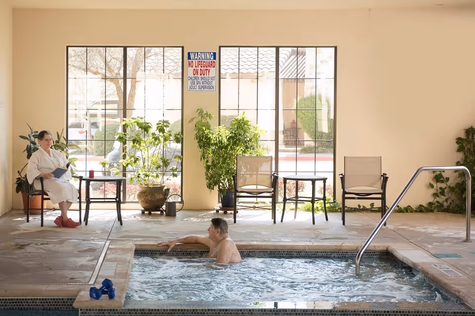 An indoor spa area with a man relaxing in a bubbling hot tub and a woman sitting nearby on a chair wearing a white robe and red slippers, holding a book. There are large windows behind them with potted plants and chairs arranged along the wall. A warning sign about no lifeguard on duty is visible between the windows.