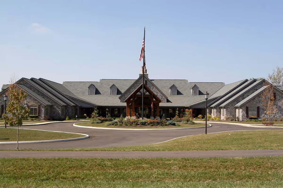 Front exterior view of The Inn at Northwood Village, a large stone building with a peaked roof and multiple dormer windows. There is a circular driveway with a landscaped island in the center featuring an American flag on a flagpole. The sky is clear and blue.