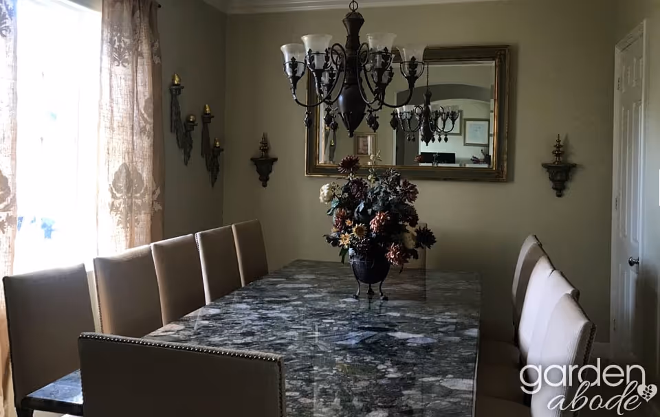 A dining room with a large rectangular marble table surrounded by beige upholstered chairs. A floral centerpiece is placed on the table. The room has beige walls with decorative wall sconces and a large mirror reflecting a chandelier hanging above the table. Light filters in through a window with patterned curtains on the left side.