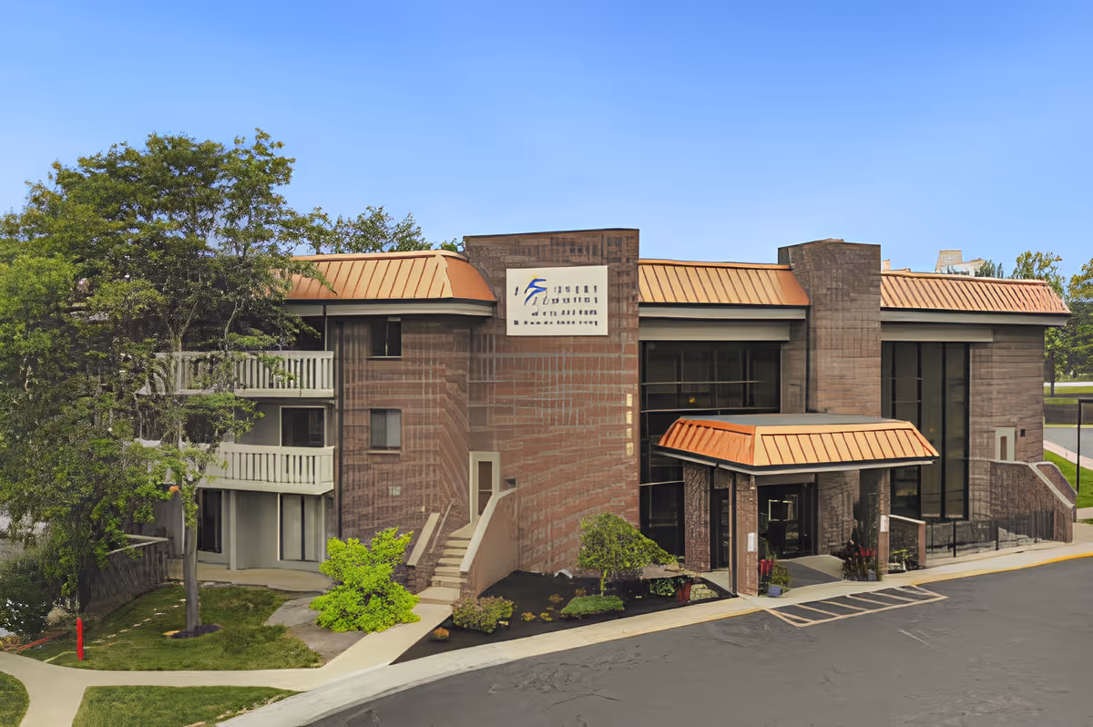 Exterior view of King's Reserve College Park building with a brick facade, large windows, and a copper-colored roof. There are trees and shrubs around the building and a paved driveway in front.