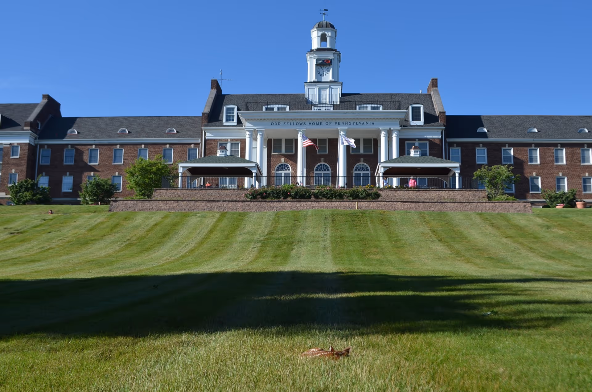 Front exterior view of a large brick building with white columns and a clock tower, labeled 'Odd Fellows Home of Pennsylvania,' with two flags flying in front and a well-maintained lawn in the foreground under a clear blue sky.