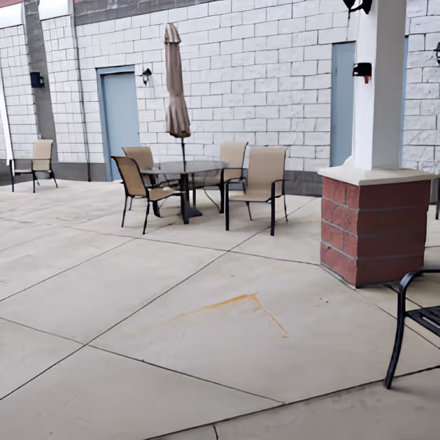 Outdoor patio area with a round glass table, four beige chairs, and a closed beige umbrella. The patio is paved with concrete and is adjacent to a building with a white brick wall and two gray doors. A red brick column supports a white overhang on the right side of the image.
