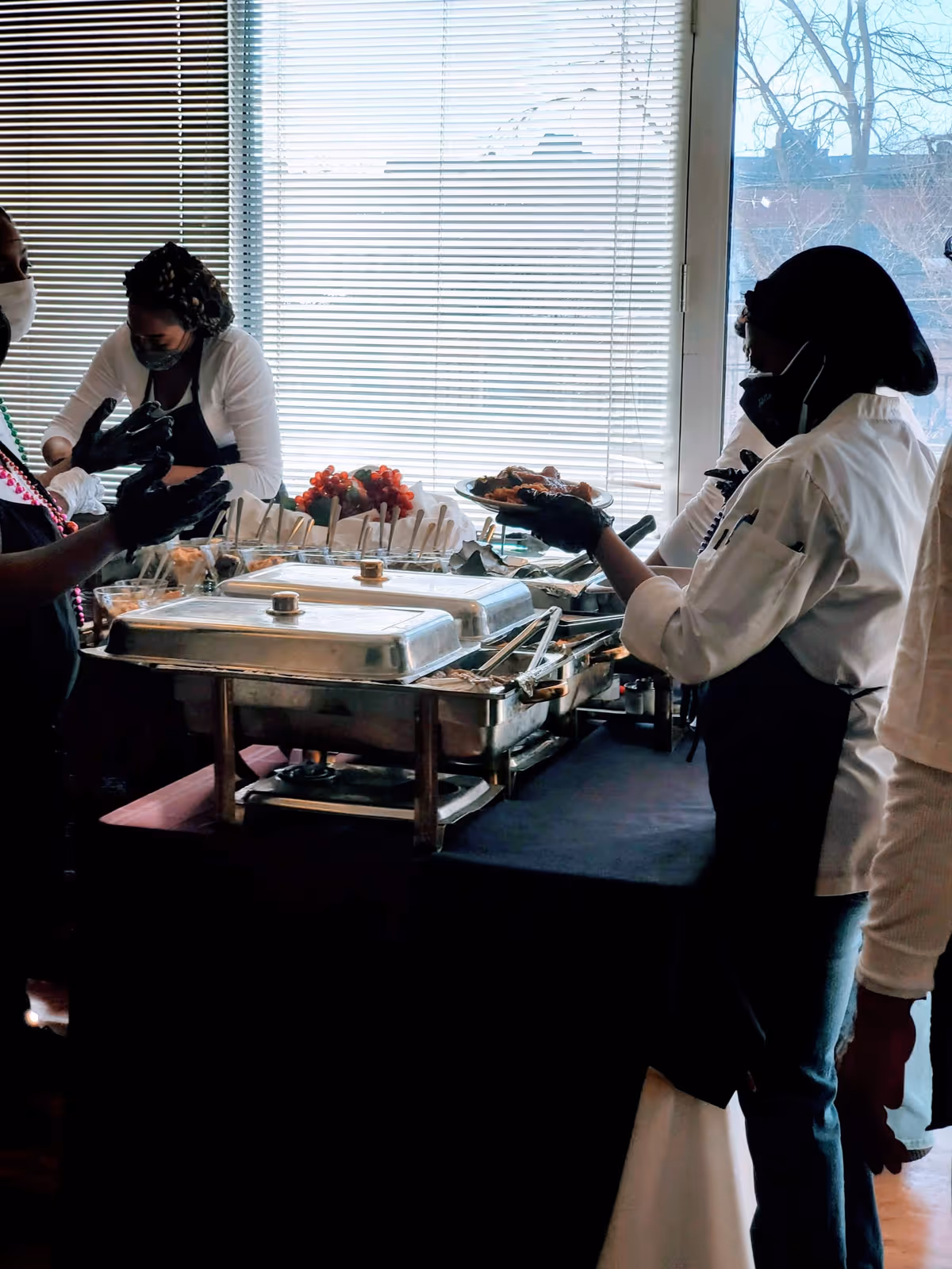 Several people wearing masks and gloves serving food from chafing dishes on a buffet table in front of a large window with blinds.