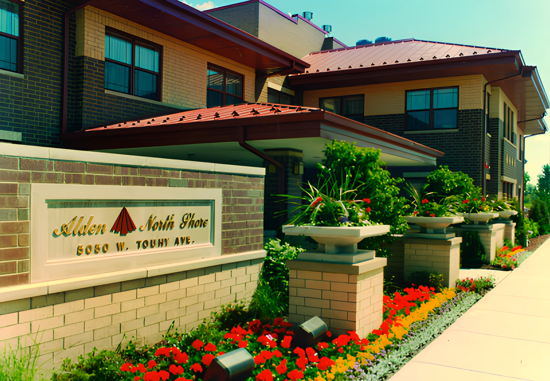 Exterior view of Alden North Shore Rehab & Health Care Center building with a brick facade, red roof, and large windows. In front of the building is a landscaped garden with colorful flowers and decorative planters along a sidewalk. A sign on a brick wall displays the facility name and address.
