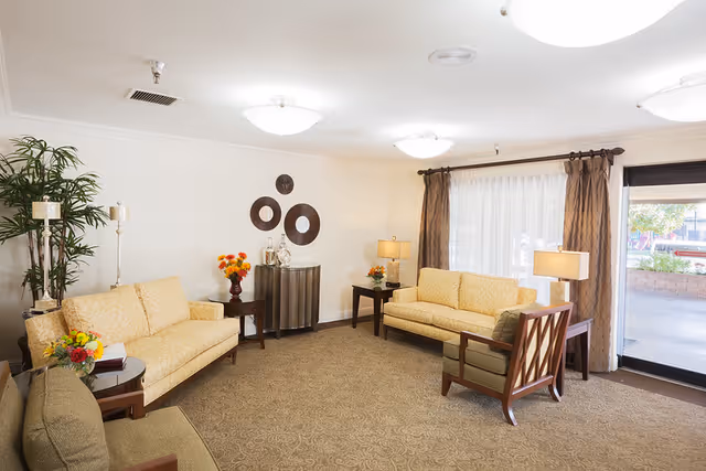 A well-lit living room with two beige sofas, a green armchair, side tables with lamps and flower arrangements, a tall potted plant, decorative wall art, and large windows with brown curtains overlooking an outdoor area.