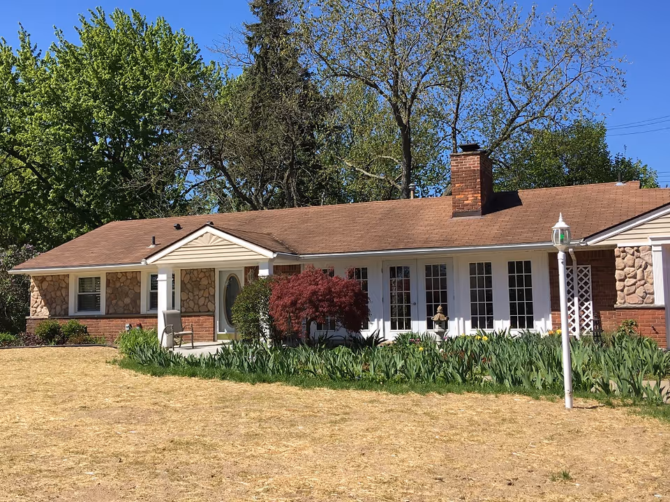 Single-story residential building with a brown shingled roof, stone and brick exterior walls, a chimney, and multiple windows. The front yard has a patchy lawn, green plants, a small red tree, and a white lamp post. Tall trees with green leaves are visible in the background under a clear blue sky.