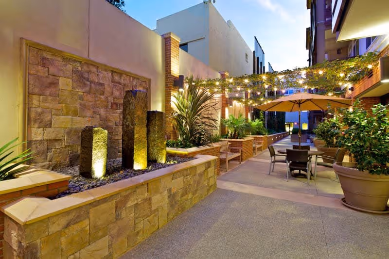 Outdoor patio area at dusk with stone water feature, potted plants, string lights overhead, tables with umbrellas, and benches along the walkway between buildings.