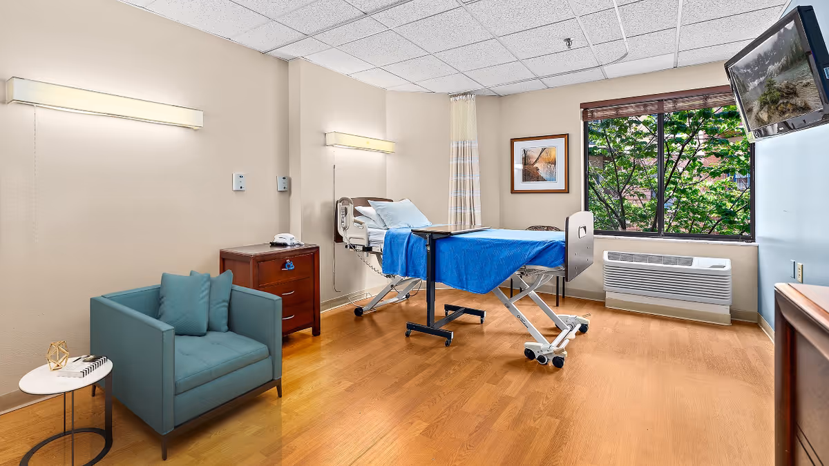 A bright and clean patient room in a skilled nursing facility featuring a hospital bed with blue bedding, a wooden nightstand with a telephone, a teal armchair with cushions, a small round side table, a large window with a view of green trees, a wall-mounted TV, and light wood flooring.