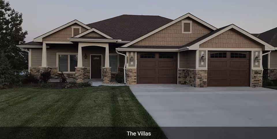 Exterior view of a single-story residential building with a front porch, two garage doors, and a well-maintained lawn. The building features beige siding with stone accents and a dark shingled roof.