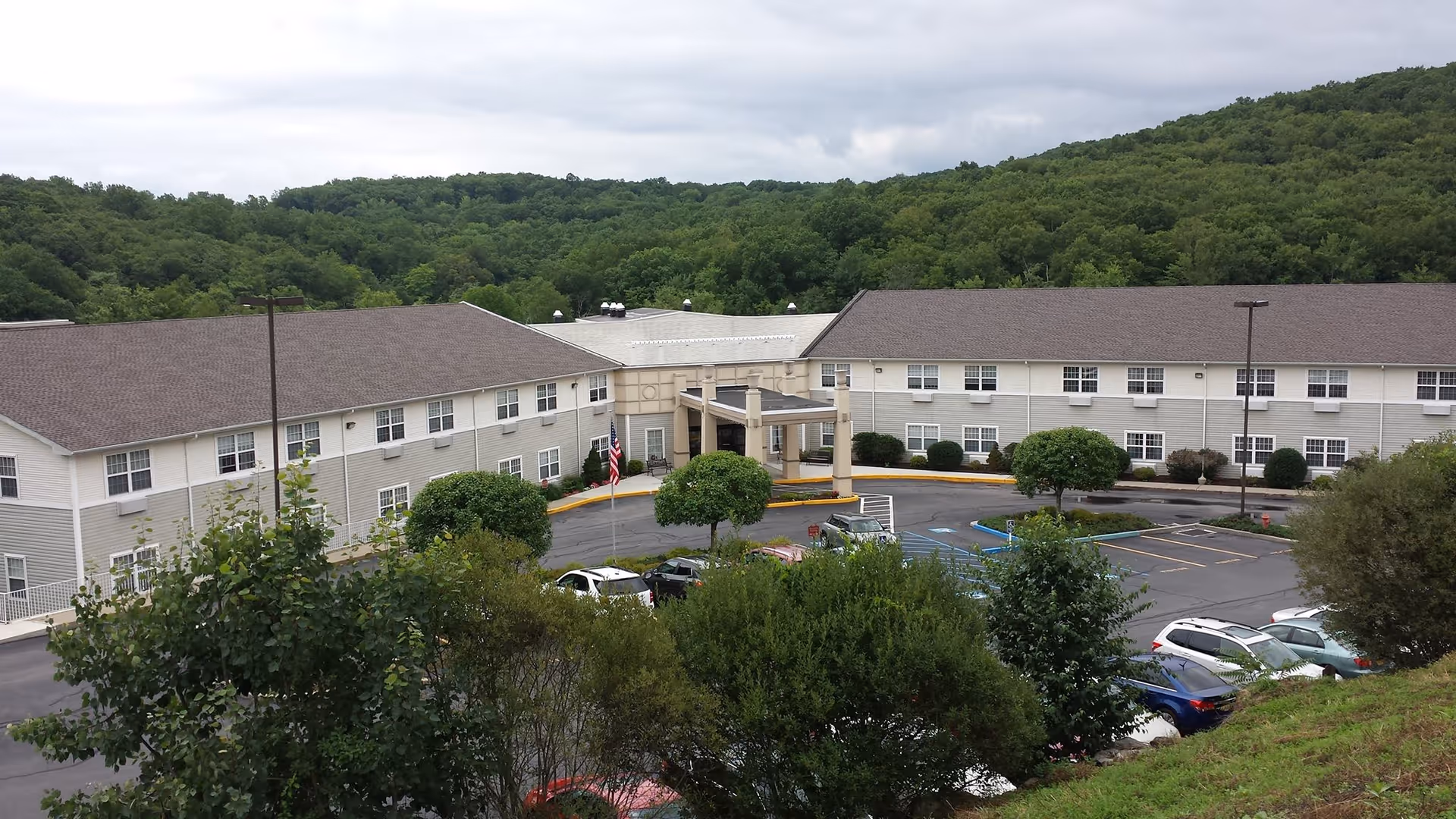 Exterior view of The Plaza at Clover Lake, a two-story senior living facility surrounded by trees and hills, with a parking lot and several cars in front.