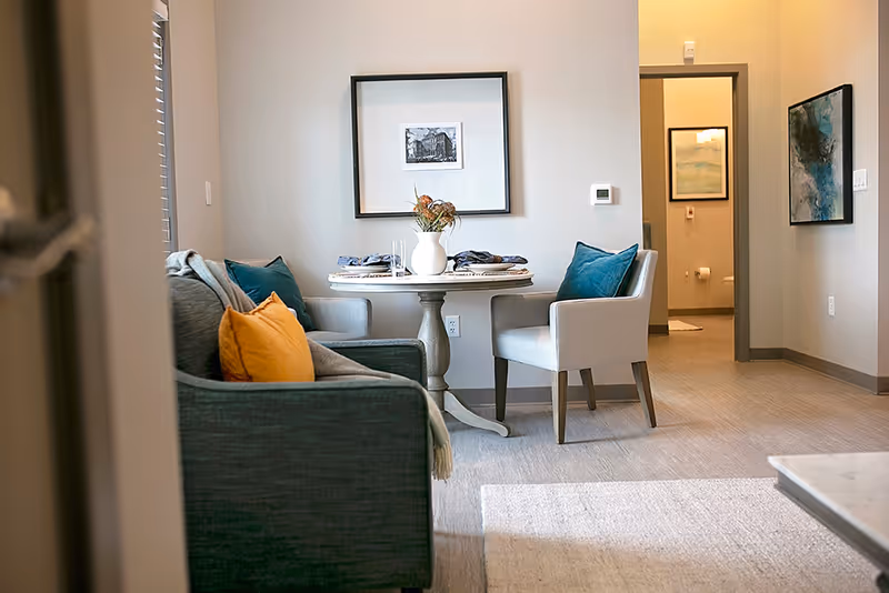 A cozy interior space featuring a small round dining table set for two with a white vase of flowers in the center. Two upholstered chairs with blue cushions are positioned around the table. To the left, a dark gray sofa with orange and blue throw pillows is partially visible. The walls are decorated with framed artwork, and an open doorway reveals a bathroom in the background.