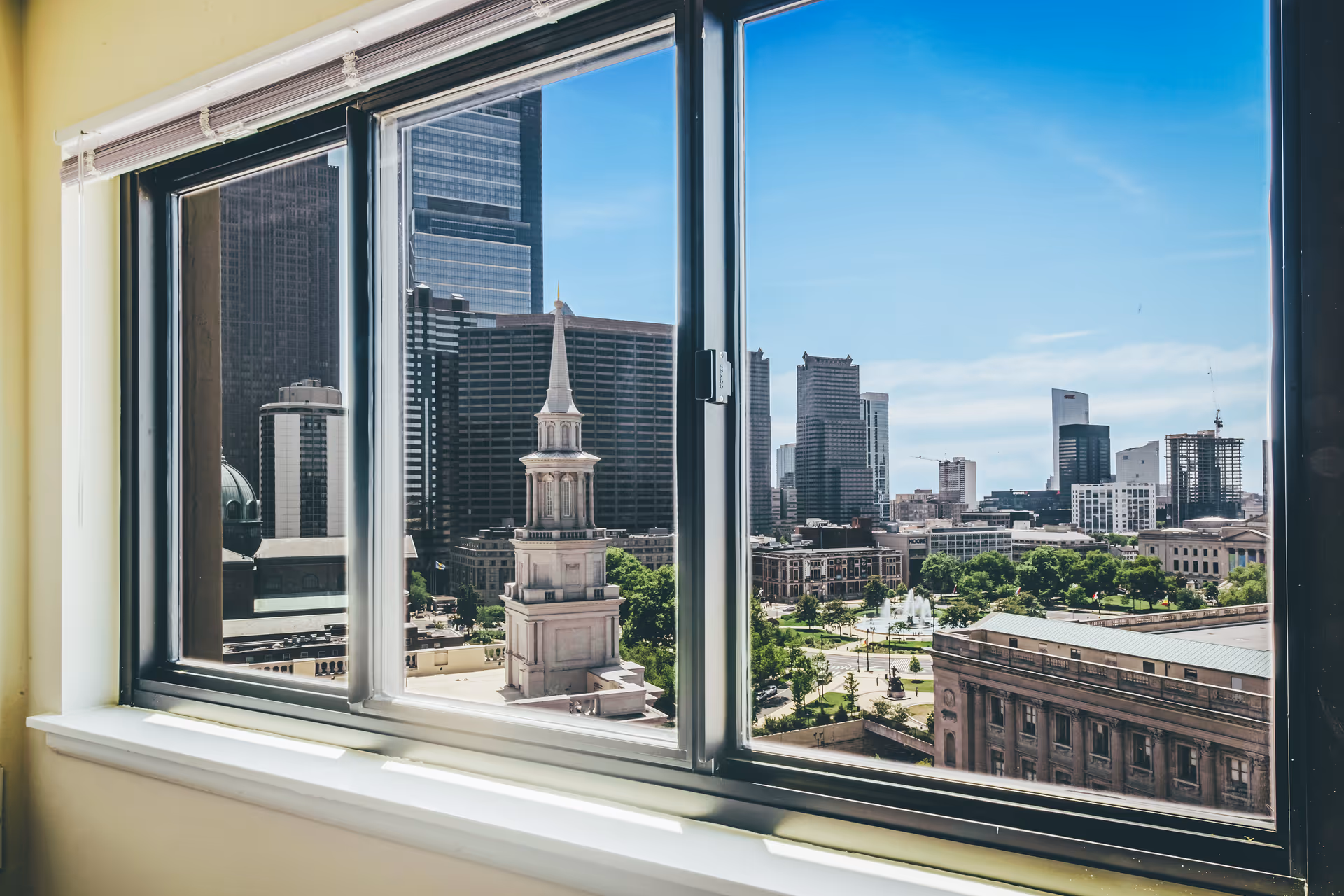 View of a cityscape through a large window with a white windowsill, showing tall buildings, a church steeple, and a park with trees and a fountain under a clear blue sky.