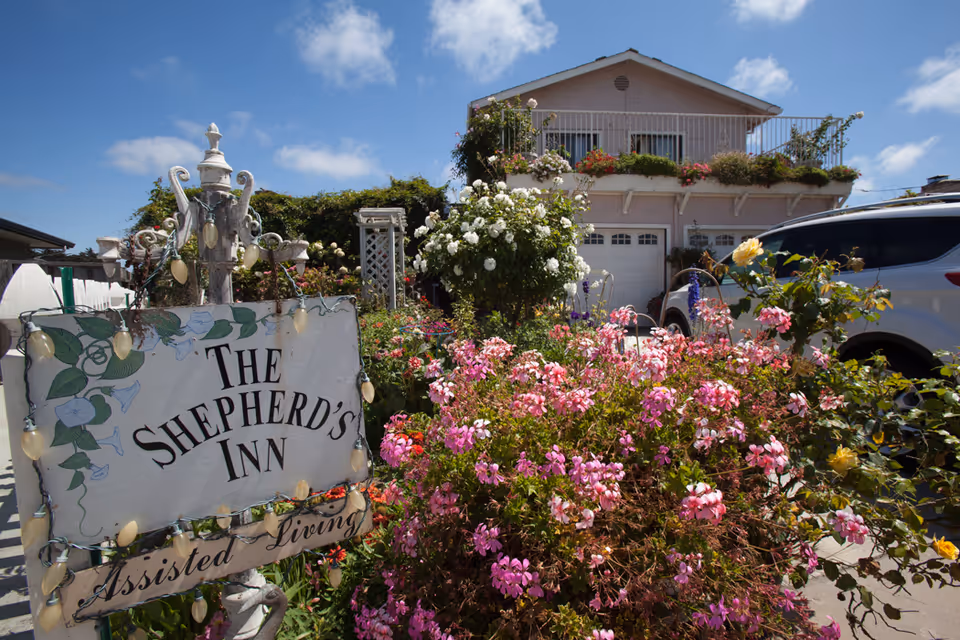 Exterior view of The Shepherd's Inn Assisted Living facility with a garden full of colorful flowers in the foreground, a white sign with the facility's name, and a two-story building with balconies and garage doors in the background under a blue sky with some clouds.