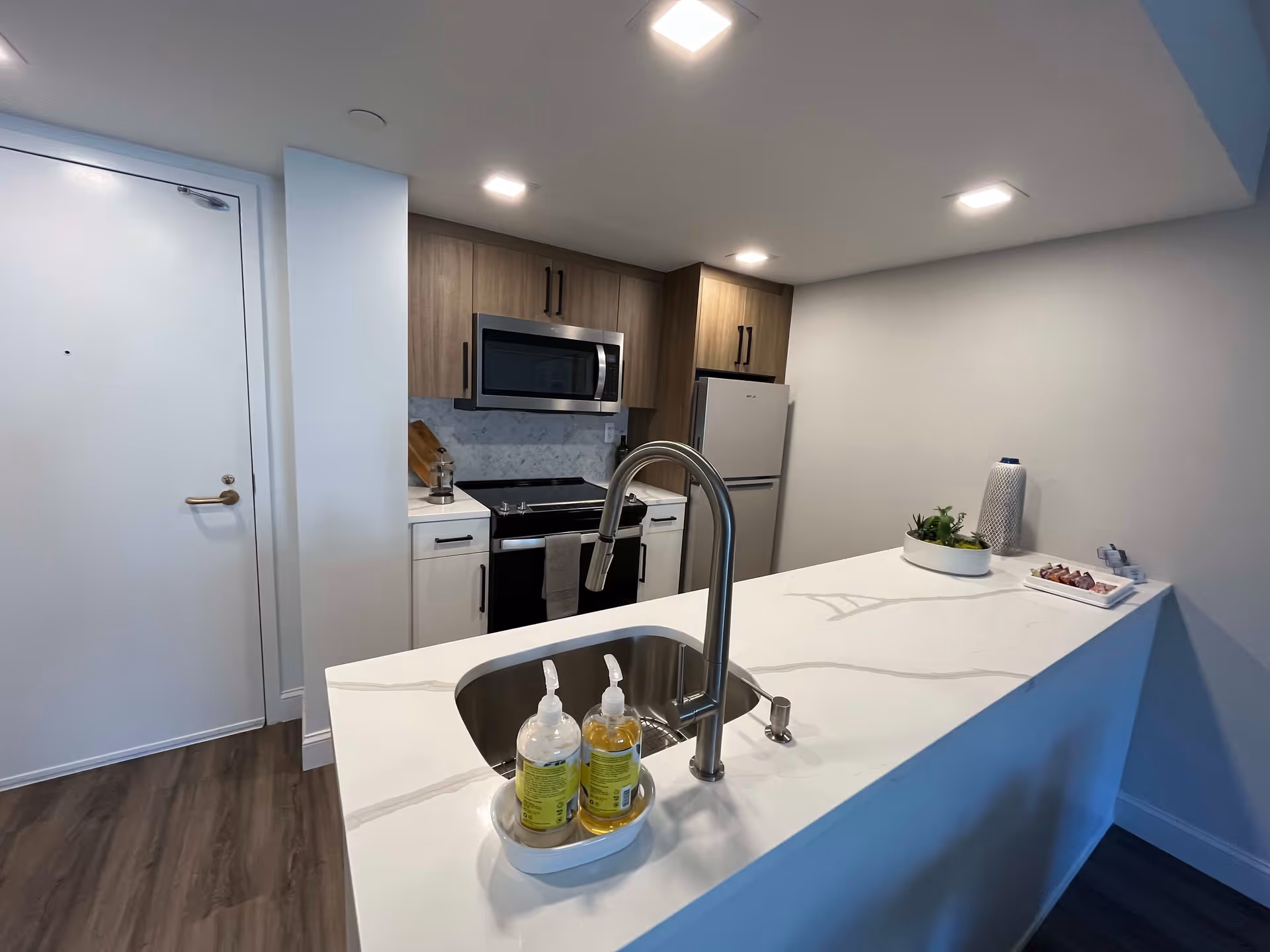 Modern kitchen area with a white marble countertop island featuring a sink and two soap dispensers. The kitchen has wooden cabinets, a stainless steel microwave, stove, and refrigerator. The floor is wood, and the walls are painted white.