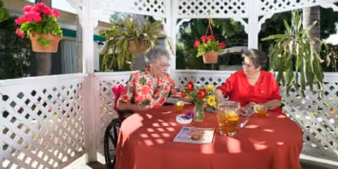 Two elderly women sitting at a round table with a red tablecloth under a white lattice gazebo. They are enjoying drinks and flowers are hanging and placed on the table. The setting is outdoors with greenery visible in the background.