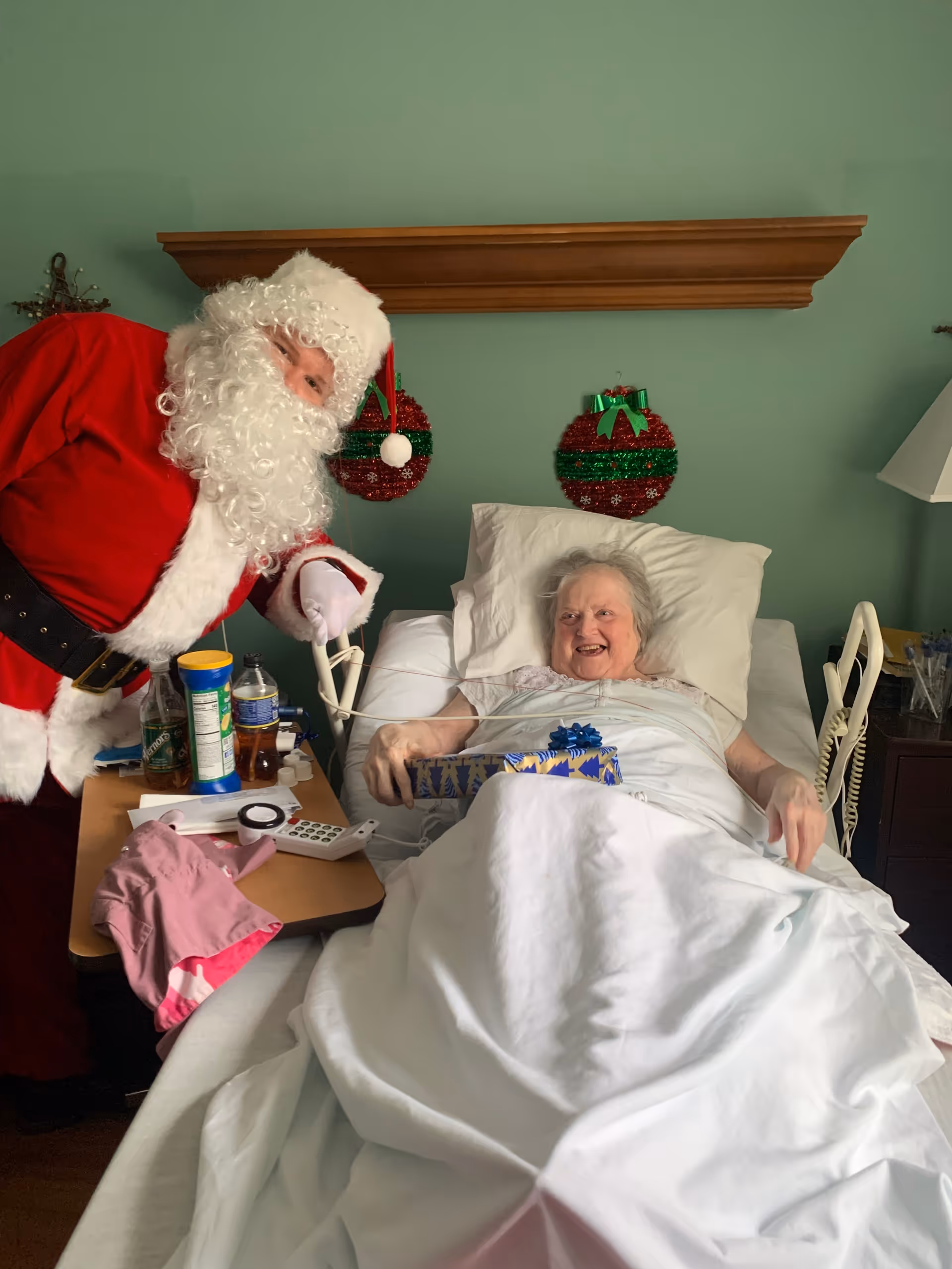 An elderly woman lying in a hospital bed covered with a white blanket, smiling and holding a wrapped gift with a blue bow. A person dressed as Santa Claus is leaning over beside her, also smiling. The room has green walls with two red and green Christmas wreath decorations hanging behind the bed. A bedside table with various items including a water bottle, snacks, and a remote control is next to the bed.