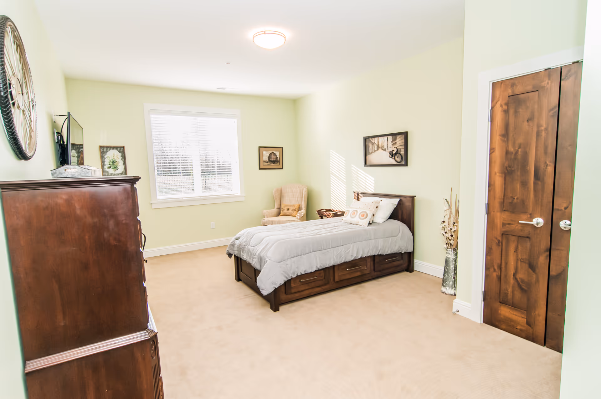 A bright and clean bedroom with light green walls and beige carpet. The room features a single wooden bed with storage drawers underneath, a beige armchair in the corner, a wooden dresser with a decorative clock and framed pictures on top, and a wooden double-door closet. Sunlight streams in through a window with white blinds.