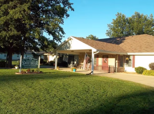 Exterior view of Sweetbriar Retirement Home showing a single-story building with a covered porch, rocking chairs, an American flag, and a well-maintained lawn with trees and a sign that reads Sweetbriar Senior Living.