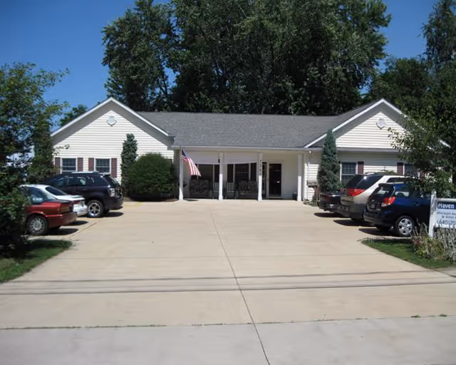Front exterior view of a single-story assisted living facility building with a gray roof and white siding. There is a concrete driveway and parking area with several parked cars on both sides. The entrance has a covered porch with columns and an American flag hanging near the door. Trees and bushes surround the building under a clear blue sky.