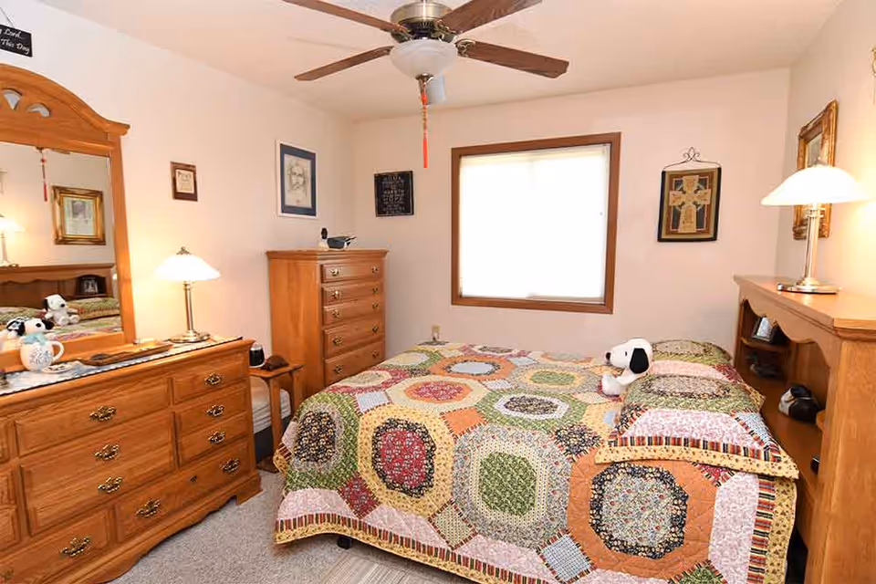 A cozy bedroom with a colorful patchwork quilt on the bed, a wooden dresser with a mirror, a tall chest of drawers, and a ceiling fan. The room has a window with a blind, framed artwork on the walls, and two bedside lamps providing warm lighting. A small Snoopy stuffed toy is placed on the bed.