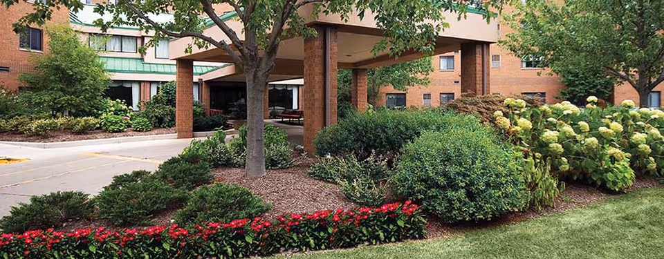 Entrance area of Ohio Living Swan Creek facility showing a covered drop-off with brick pillars, surrounded by well-maintained landscaping including bushes, trees, and flower beds with red and white flowers.