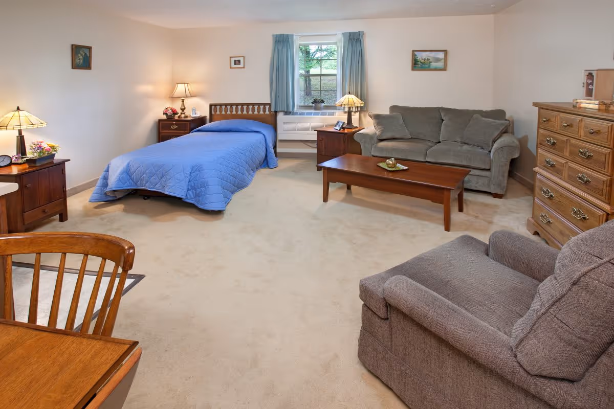 A cozy senior living room with a single bed covered in a blue quilt, a gray sofa with cushions, a wooden coffee table, a wooden dresser, a gray armchair, and two bedside tables with lamps. There is a window with blue curtains letting in natural light and a small plant on the windowsill. The room has beige carpet and light-colored walls with a few small framed pictures.