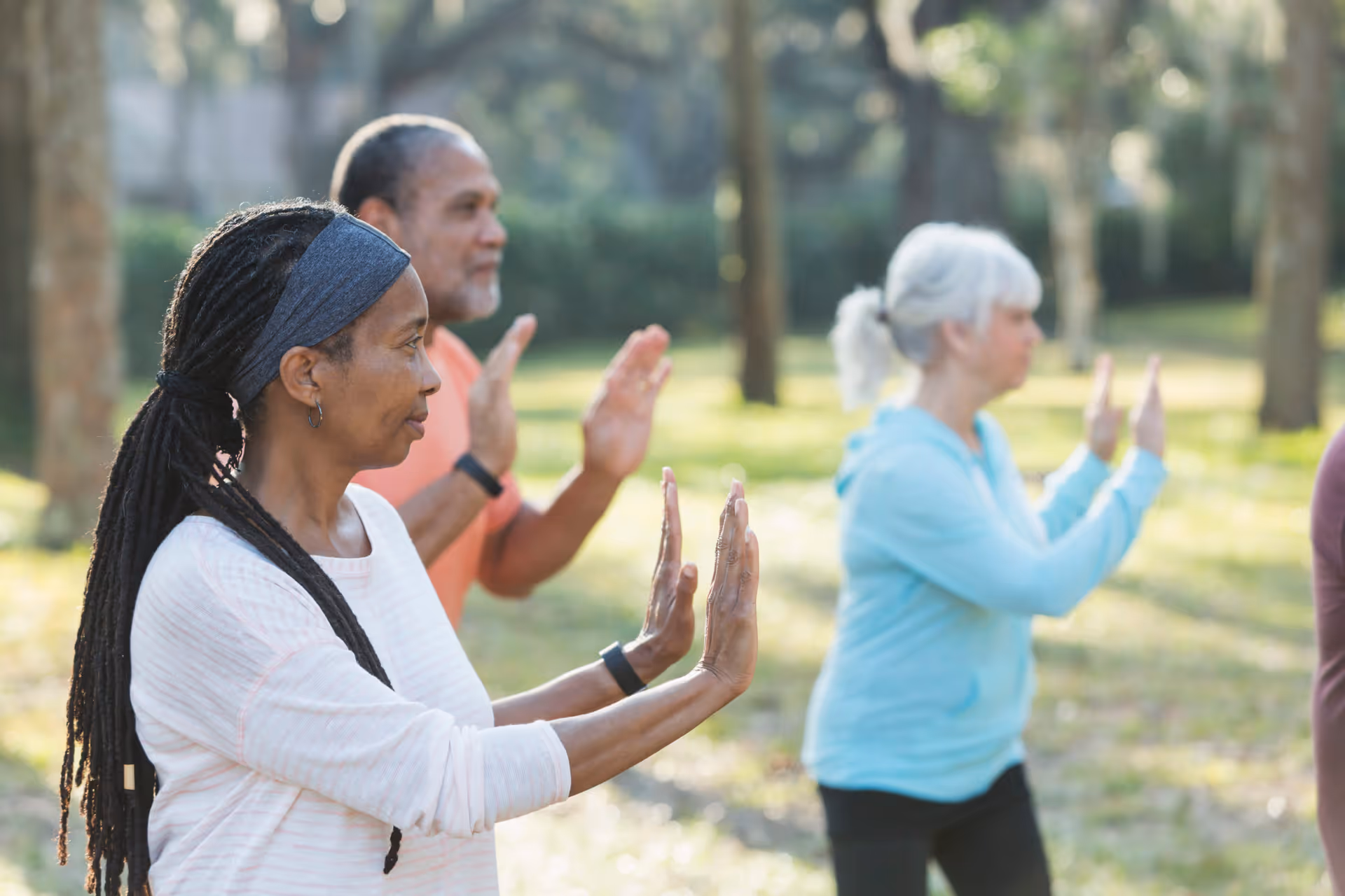 A group of senior adults practicing Tai Chi outdoors in a park-like setting with trees and grass, focusing on slow, controlled hand movements.