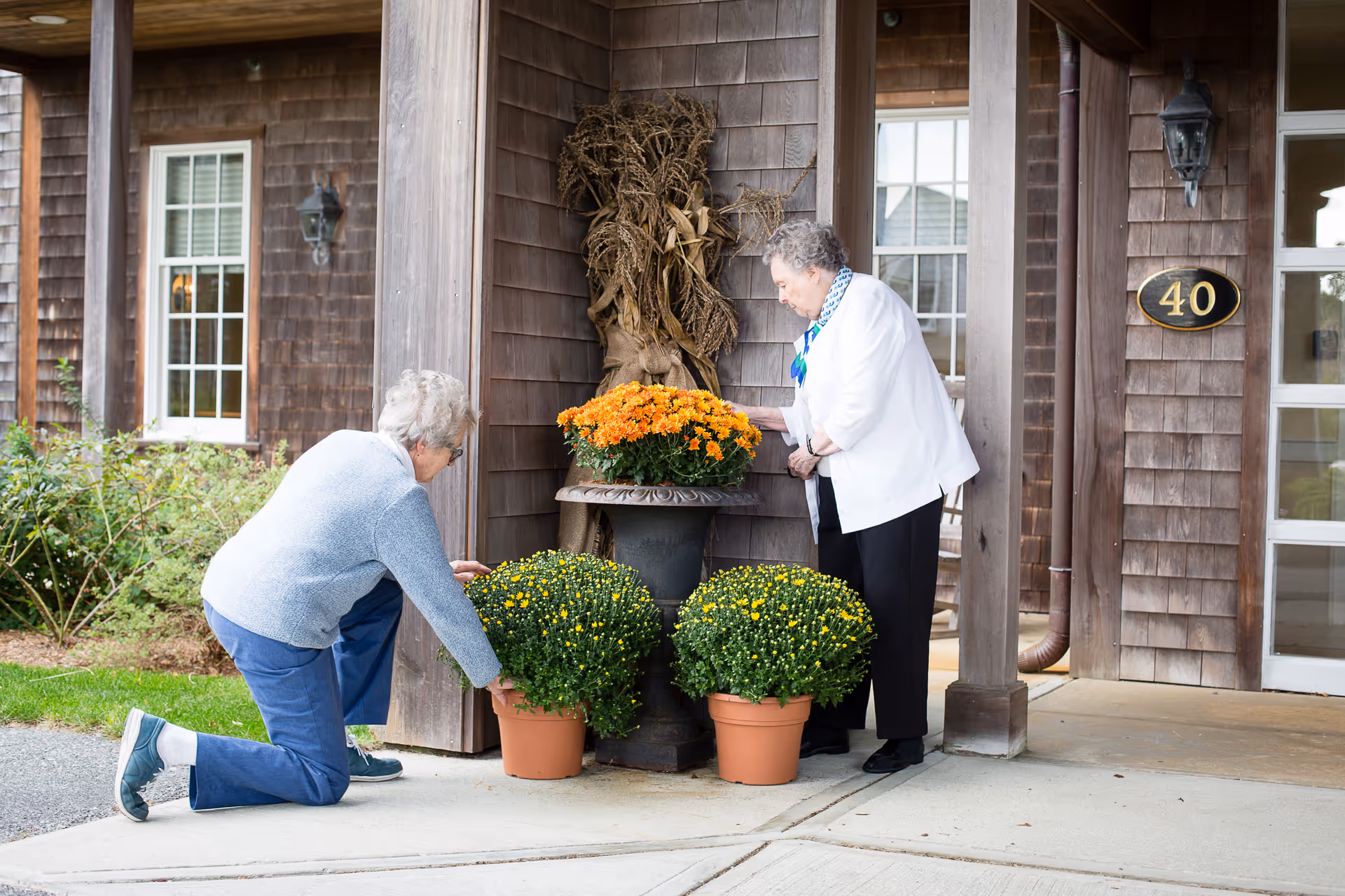 Two elderly women tending to potted flowers outside a building with wooden siding. One woman is kneeling adjusting a green potted plant, while the other is standing and touching an orange flowering plant on a pedestal. The building has a window, a door with the number 40, and some dried cornstalk decorations.