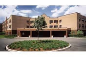 Exterior view of a multi-story brick building with a covered entrance and a circular driveway with landscaping in front under a partly cloudy sky.