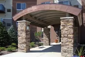 Covered entrance to a residential building with stone pillars supporting a curved roof, surrounded by landscaping including bushes and trees.