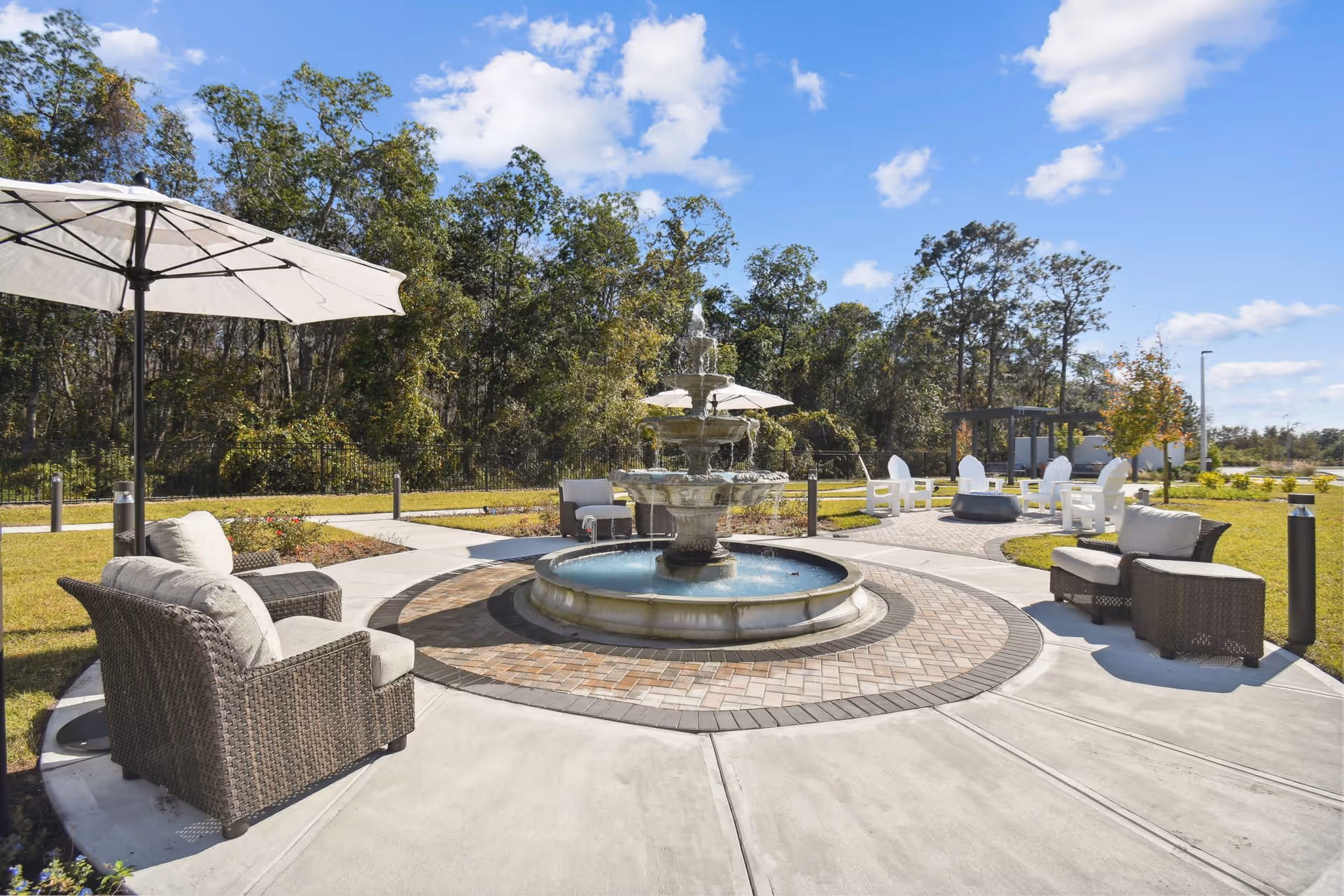 Outdoor seating area with cushioned wicker chairs and umbrellas arranged around a large tiered water fountain, surrounded by a paved circular walkway and green lawn with trees in the background under a blue sky with scattered clouds.