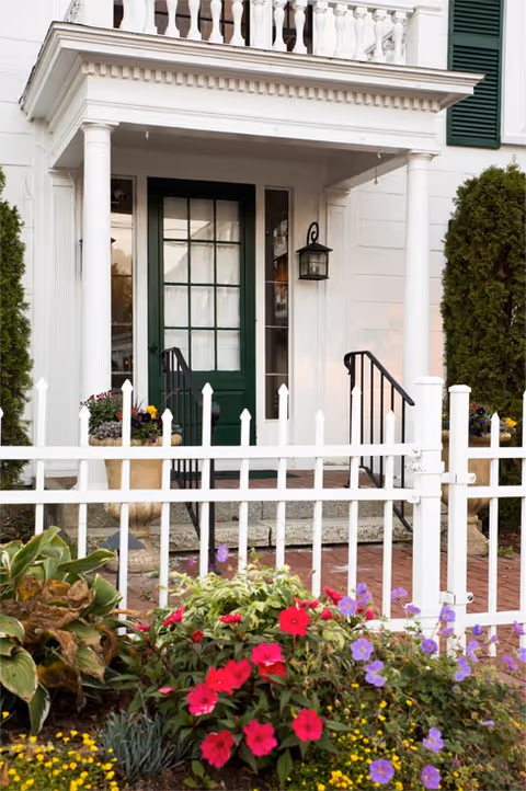 Entrance of a white building with a green door, white columns, and a small porch with black railings. A white picket fence surrounds a garden with colorful flowers including red, purple, and yellow blooms.