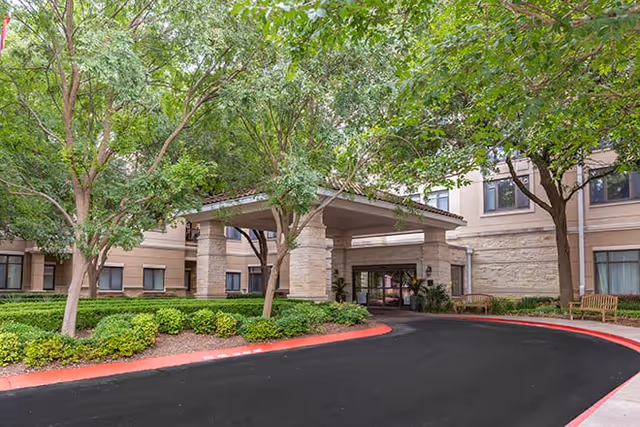Entrance of Brookdale Gaines Ranch facility with a covered driveway, surrounded by green trees and shrubs, and beige building walls with windows.