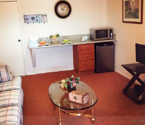 A cozy living area with a plaid sofa on the left, a glass coffee table with a floral arrangement and decorative items, a small kitchenette area with a microwave, mini fridge, and countertop with bowls and a teapot. A wall clock and framed artwork are visible on the walls.