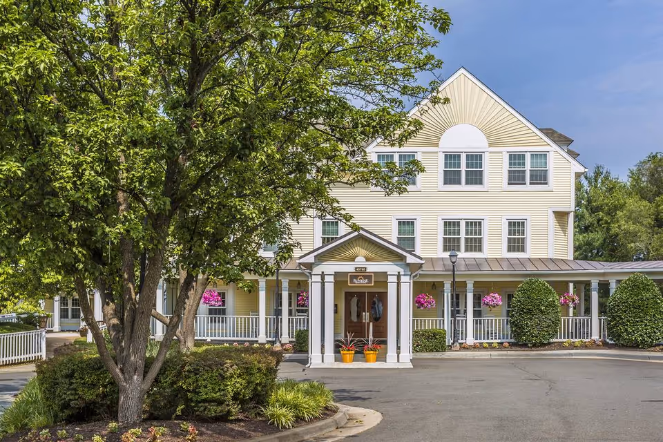 Exterior view of a three-story yellow senior living facility building with white trim and a covered entrance. The building is surrounded by greenery, including a large tree and bushes, with hanging flower baskets on the porch. The sky is clear and blue.