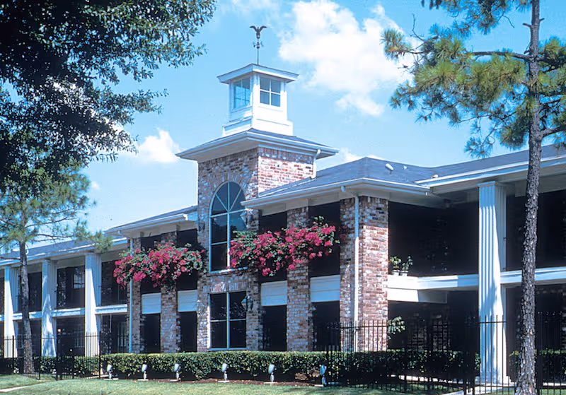 Exterior view of a two-story brick building with white columns and a cupola on the roof. The building has large windows and hanging flower baskets with pink flowers. There are trees and shrubs around the building under a blue sky with some clouds.