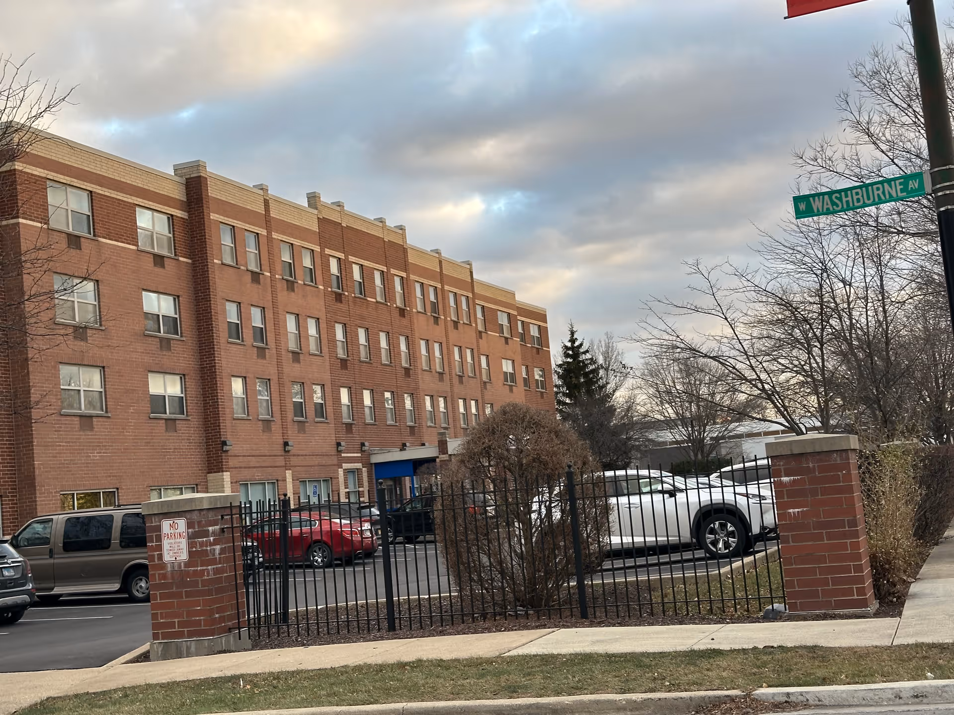 Exterior view of a multi-story brick building with multiple windows, a parking lot with several cars, a black metal fence with brick pillars, and a street sign reading W Washburne Ave under a cloudy sky.
