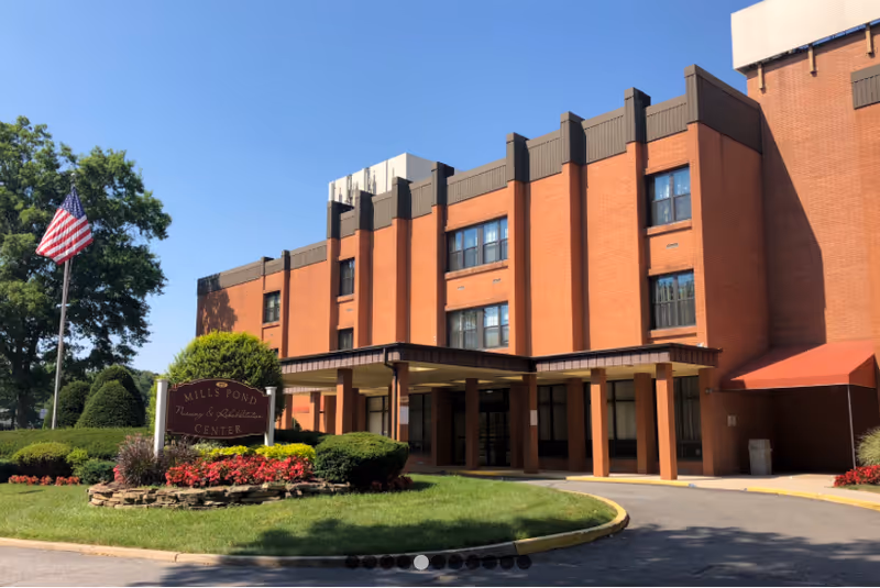 Exterior view of a multi-story brick building with a covered entrance, surrounded by well-maintained landscaping including bushes, flowers, and a lawn. An American flag is flying on a flagpole near the entrance. A sign in front reads 'Mills Pond Nursing & Rehabilitation Center'.