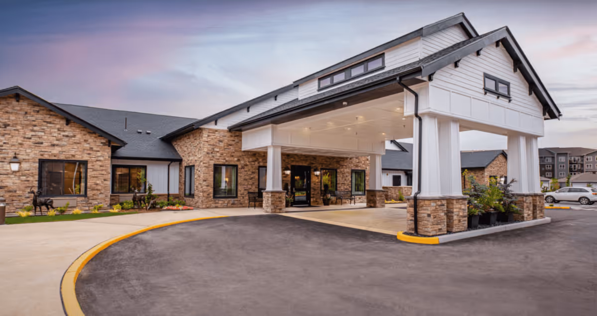 Exterior view of Deer Ridge Memory Care facility showing a covered entrance with stone and white paneling, a curved driveway, and landscaped areas with plants and decorative statues under a twilight sky.