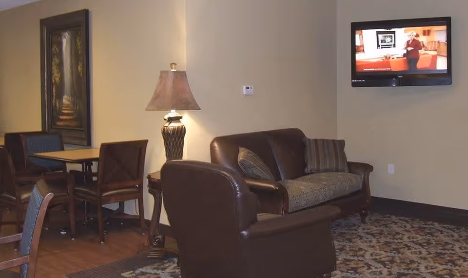 A cozy senior living common area with a brown leather armchair and a matching sofa with patterned cushions. A table lamp with a decorative base is on a side table between the seating. In the background, there is a dining table with chairs and a framed painting on the wall. A flat-screen TV is mounted on the wall showing a program.