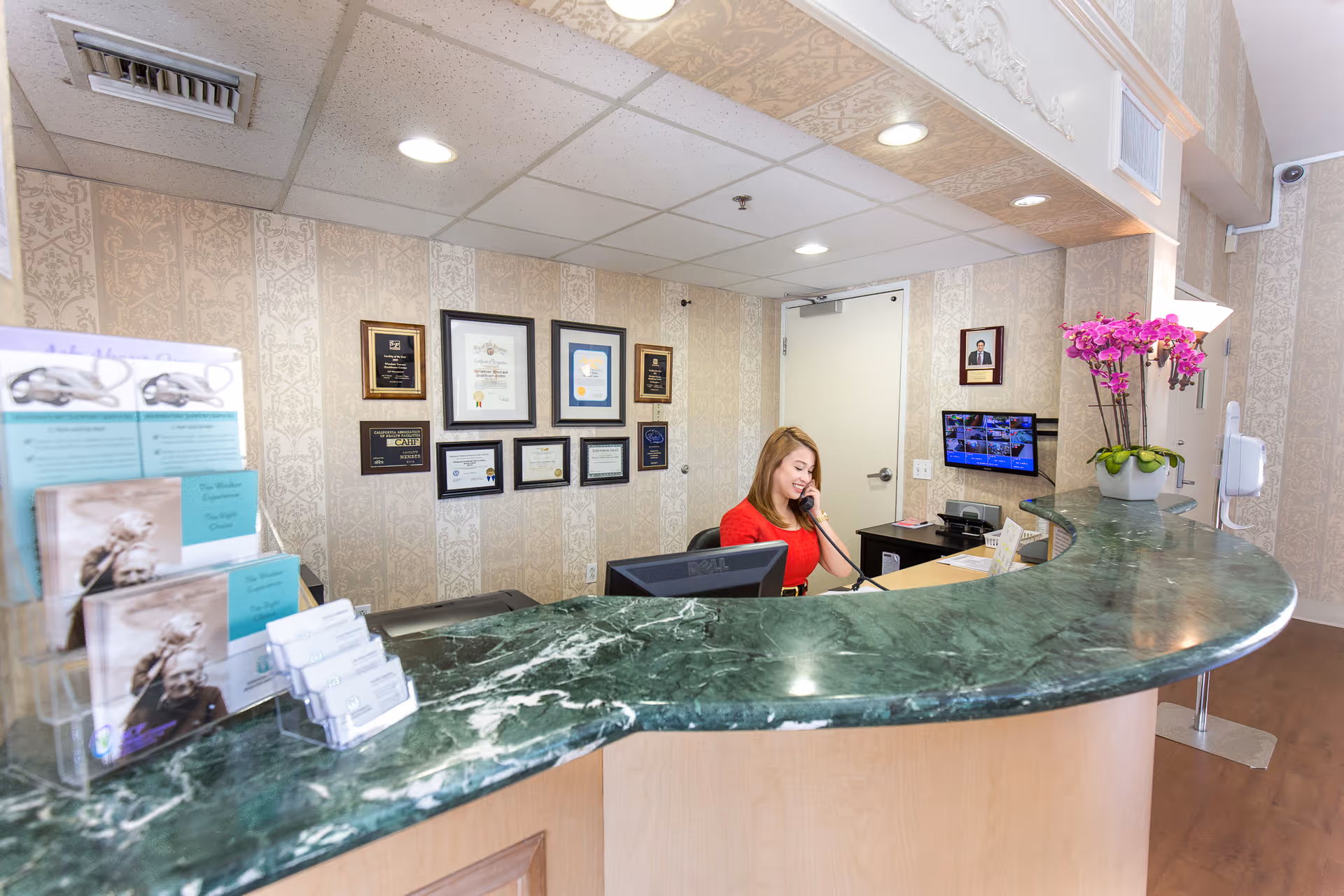 Reception area of Windsor Terrace Healthcare Center with a woman in a red dress sitting behind a green marble counter, talking on the phone. The wall behind her displays several framed certificates and awards. There is a small monitor showing security camera footage and a purple orchid plant on the counter.