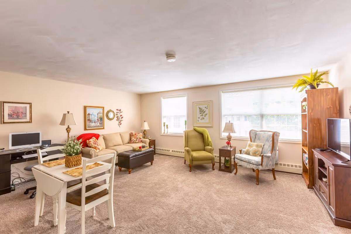 A bright and cozy living room with beige walls and carpeted floor. The room features a beige sofa with colorful cushions, two armchairs (one green and one patterned), a small wooden side table with a lamp, a wooden TV stand with a flat-screen TV, and a wooden bookshelf with a plant on top. There is also a small white dining table with two chairs and a computer desk with a monitor and keyboard. The room is decorated with framed pictures and has two large windows letting in natural light.